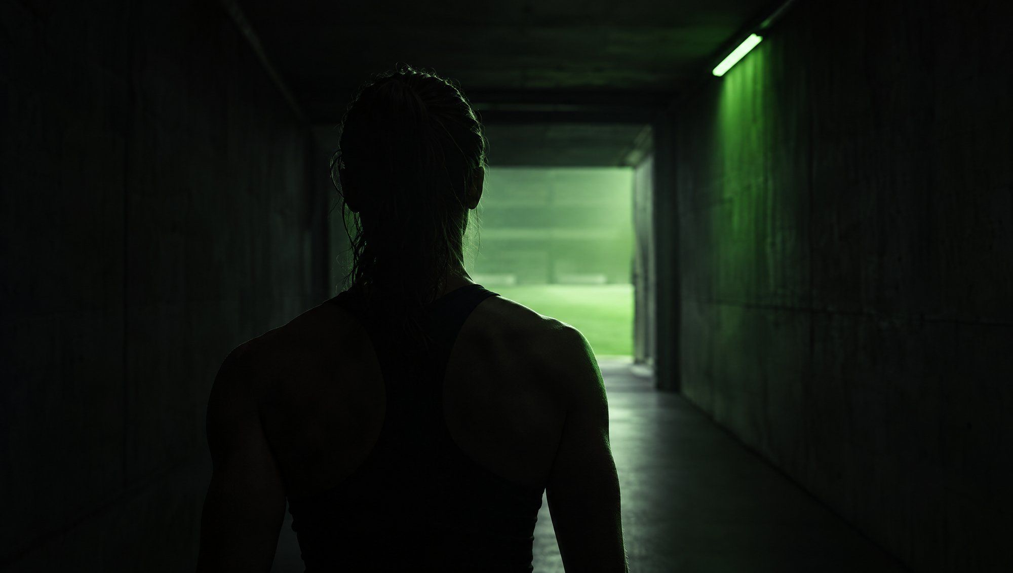 An athlete in a dark tank top, seen from behind in silhouette, walking down a dark concrete tunnel toward a green-lit field, with a green strip light on the wall.